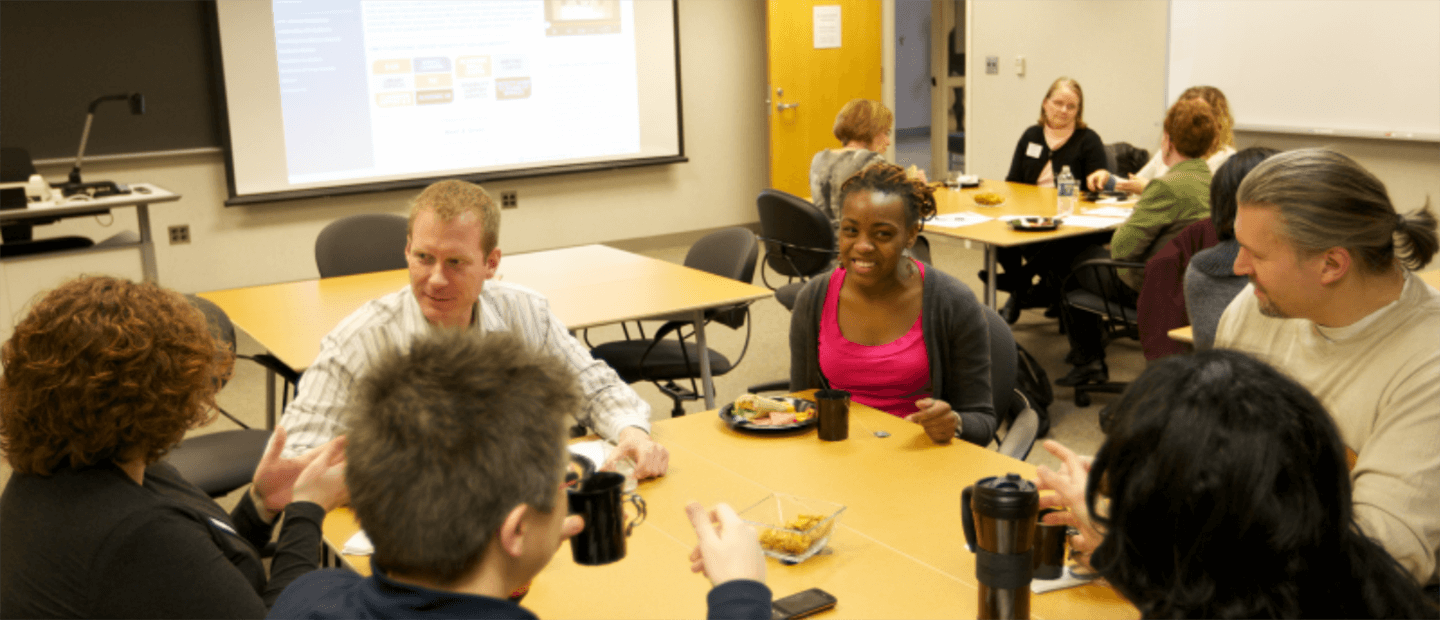 Adults seated at a two square tables in a classroom, talking and having breakfast.