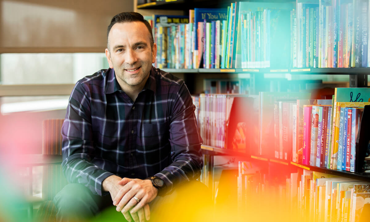 Man looks toward camera smiling. There is a library bookshelf behind him.