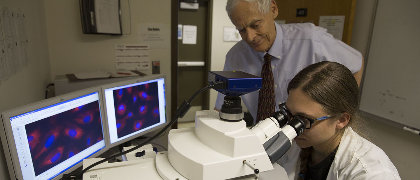 professor standing over a student looking in a microscope with two computer