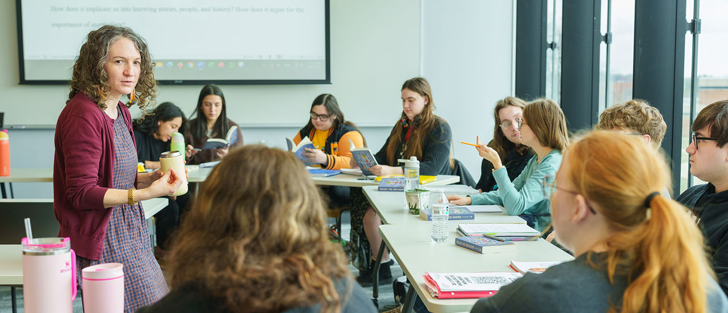 An English professor talks in front of a group of students, who are also meeting in groups.