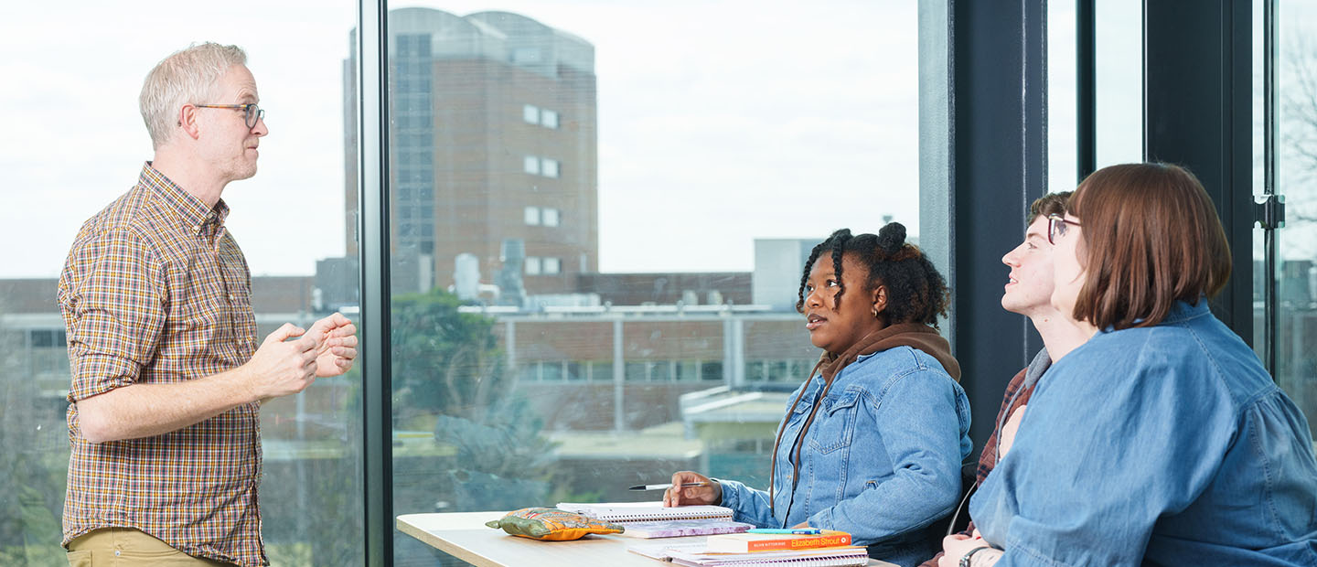A professor talks in front of three students sitting at a desk.