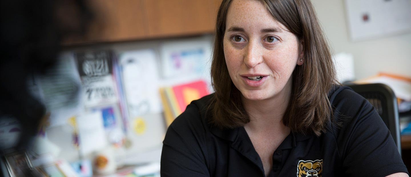 Woman in Oakland University polo shirt seated at a desk speaking to another individual who is off-camera.