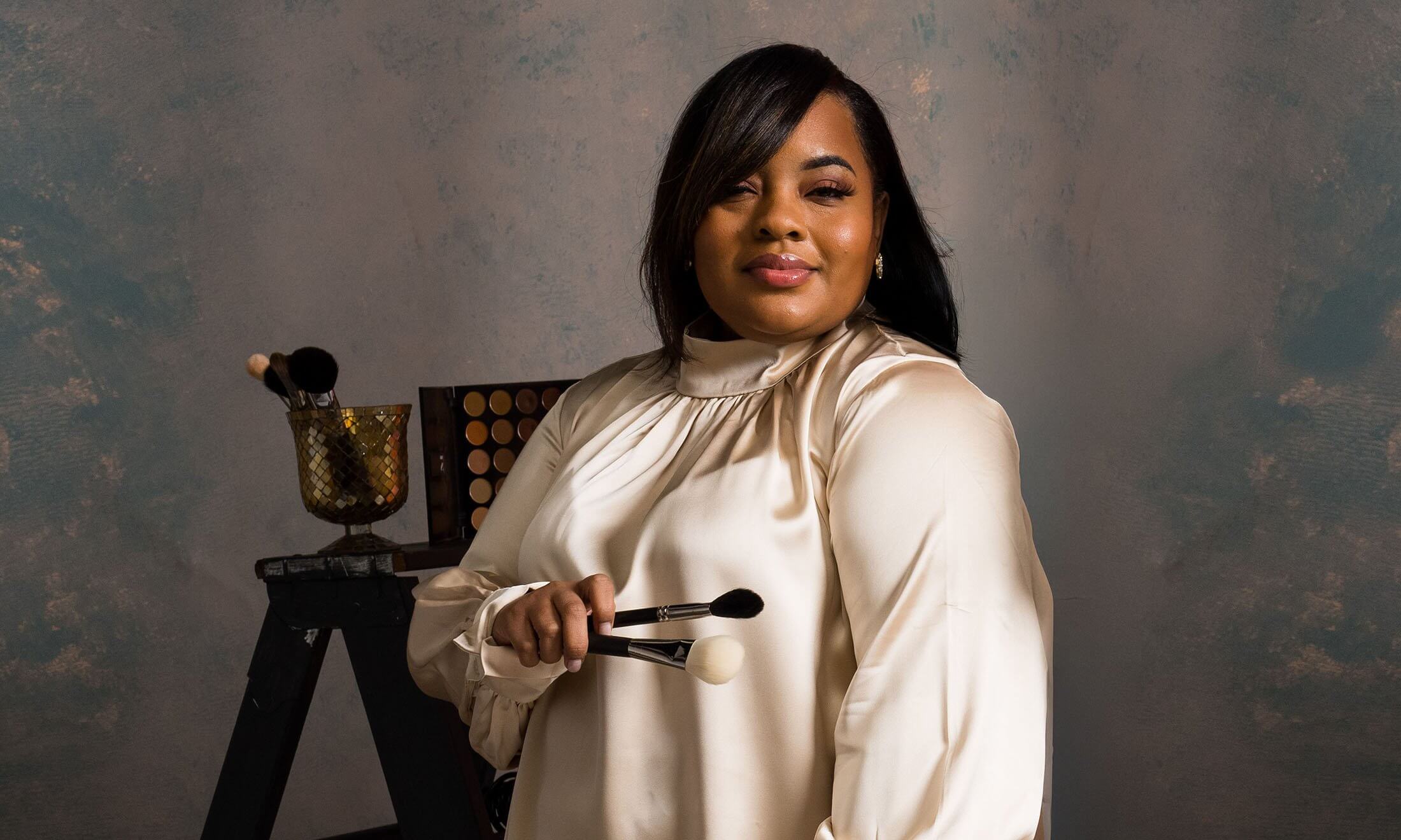 A woman holding makeup brushes looks at the camera.