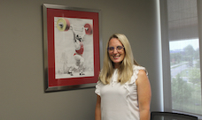 A smiling woman stands next to artwork hanging on a wall.