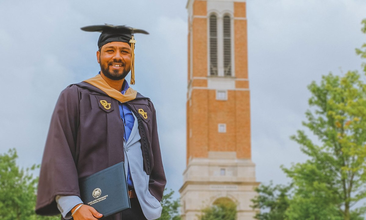 Man in cap and gown outside near brick bell tower.