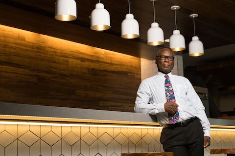 Henry Aigbedo, Ph.D., leaning on a railing in a dimly lit room.