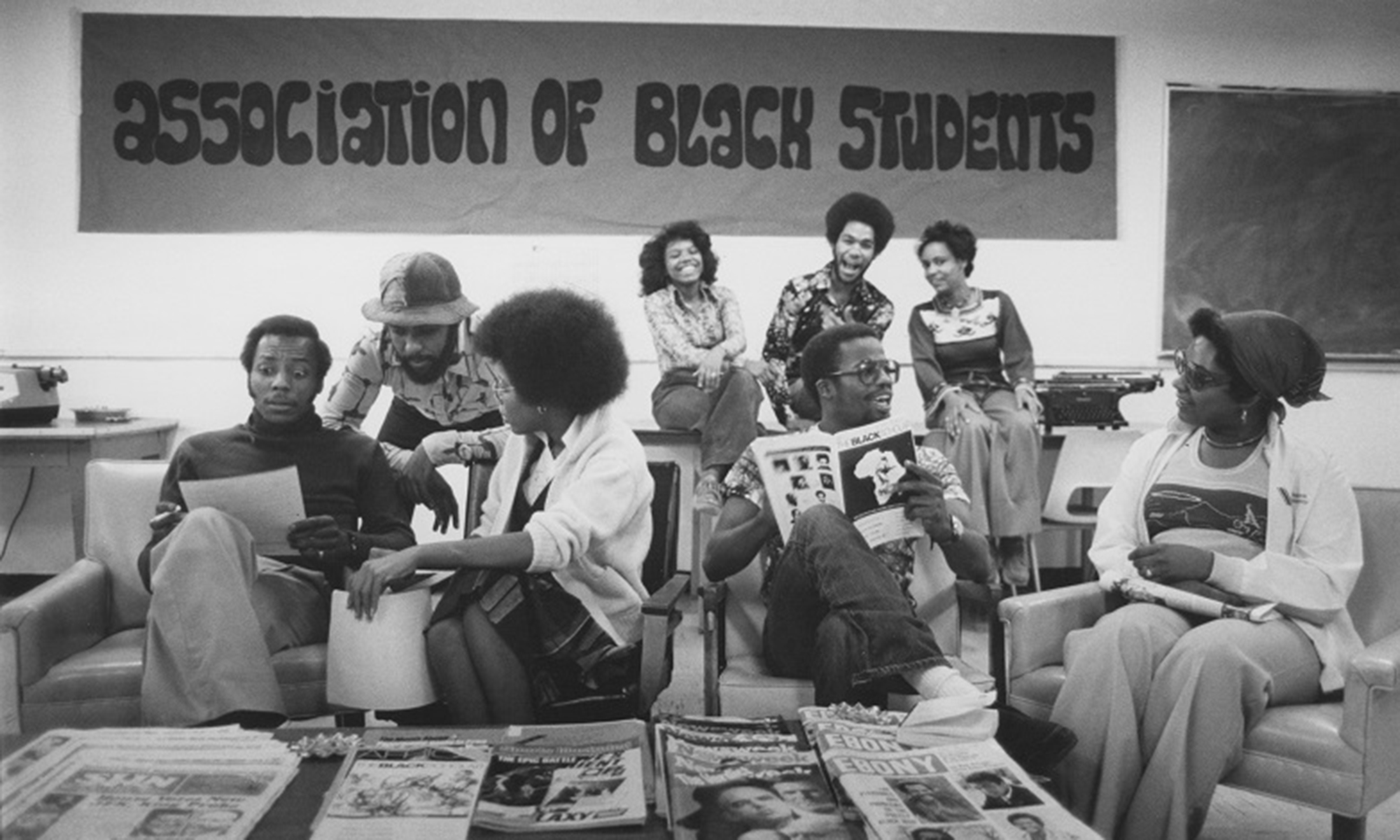 A group of students sitting around on couches, tables and chairs in front of a sign that reads, 
