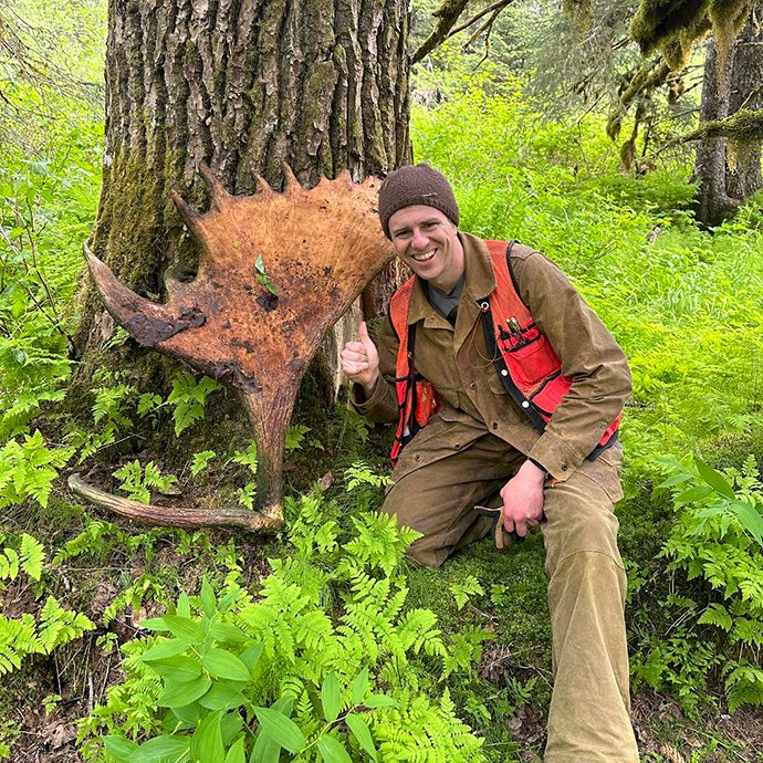 Person psoing next to some moose antlers next to a tree