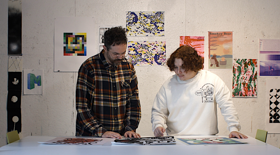 A student and professor look at prints at a large table. There is digital artwork hanging on the wall behind them.