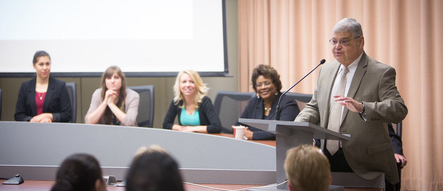 A man speaking into a microphone at a podium, with a panel of people seated behind him.