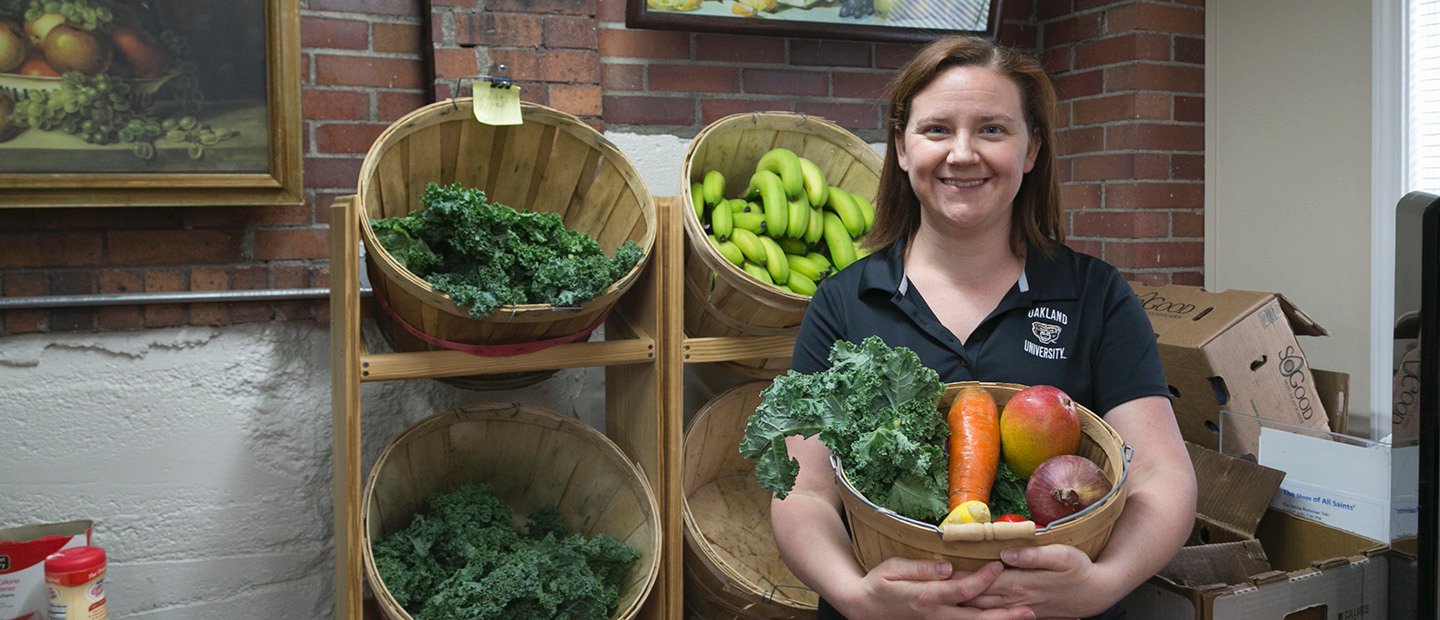 A woman holding a basket of vegetables, in front of a shelf displaying more food.