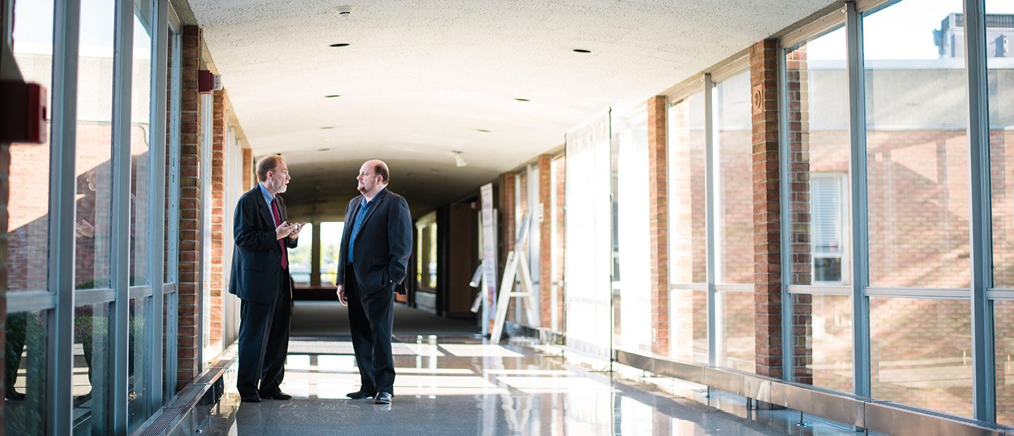 Two men having a discussion in a hallway with a lot of windows.