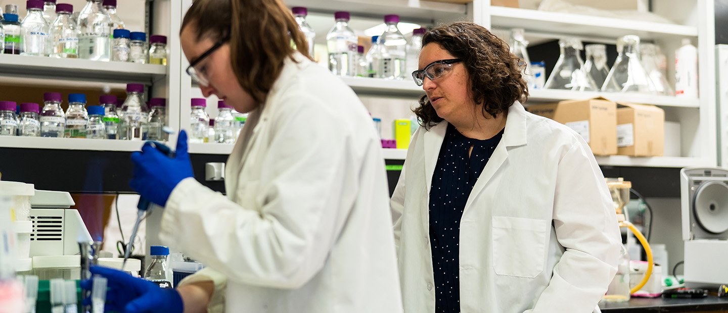 Two women in a lab with chemicals in glass jars.