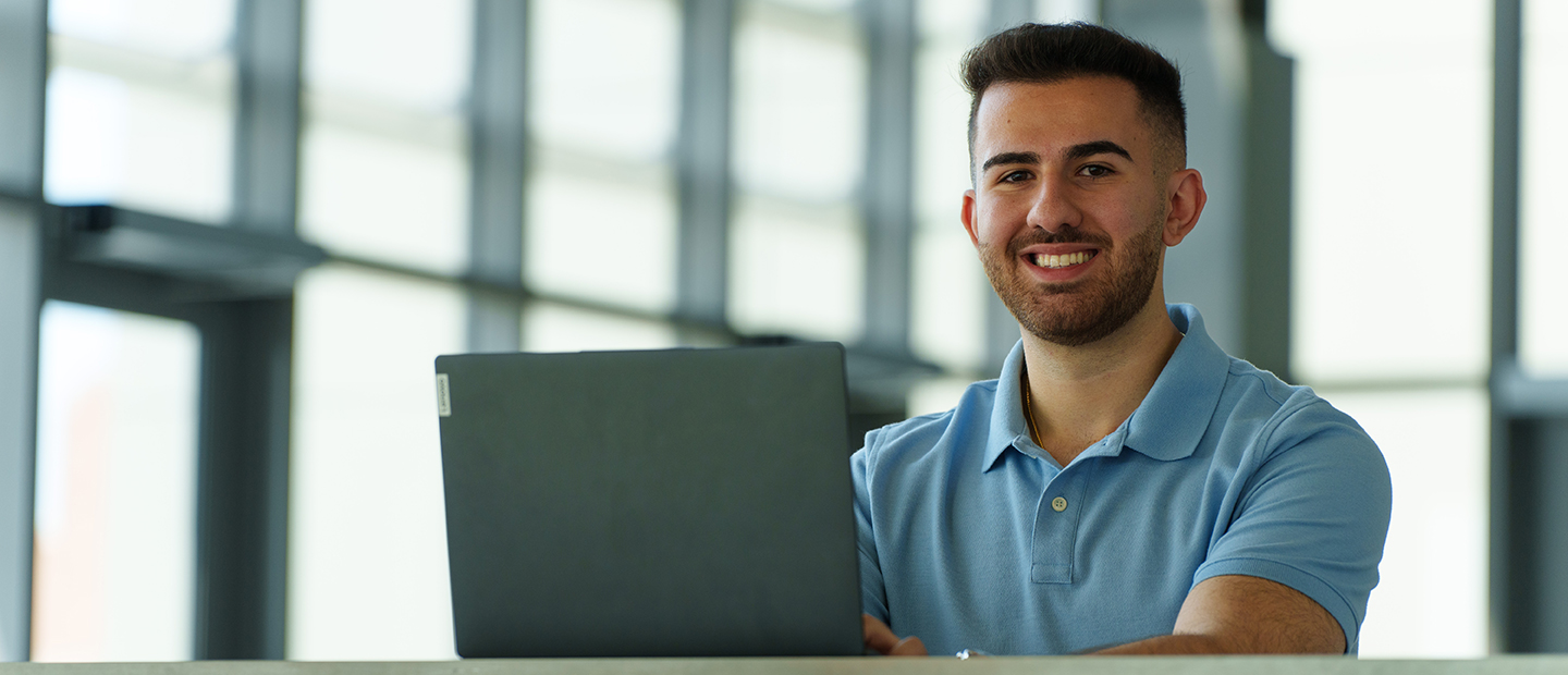 A young man with a laptop smiling at the camera