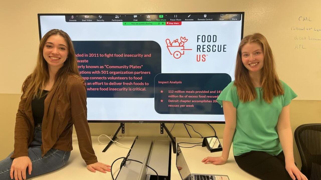 Oakland University students, Sarah Nyquist and Rachel Whalen, seated in front of a large monitor displaying information about Food Rescue US.