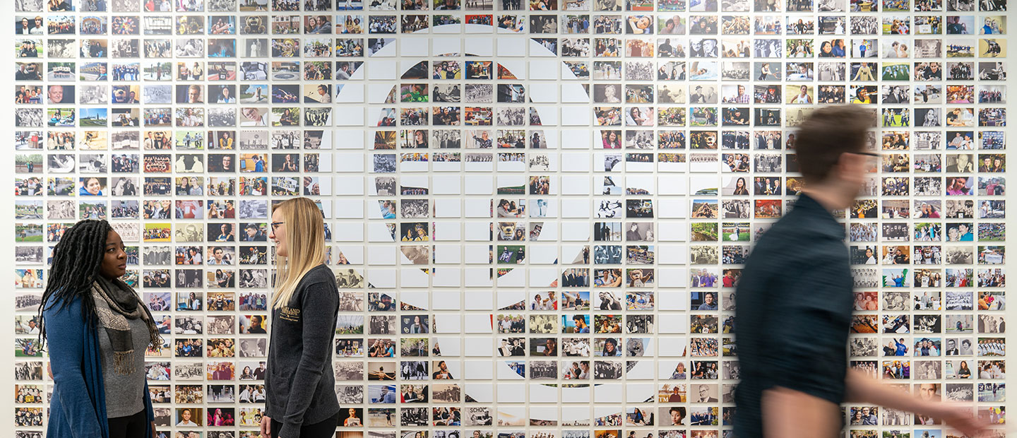 People walking by a wall with a mosaic of photo tiles and the interlocking O U in the middle.