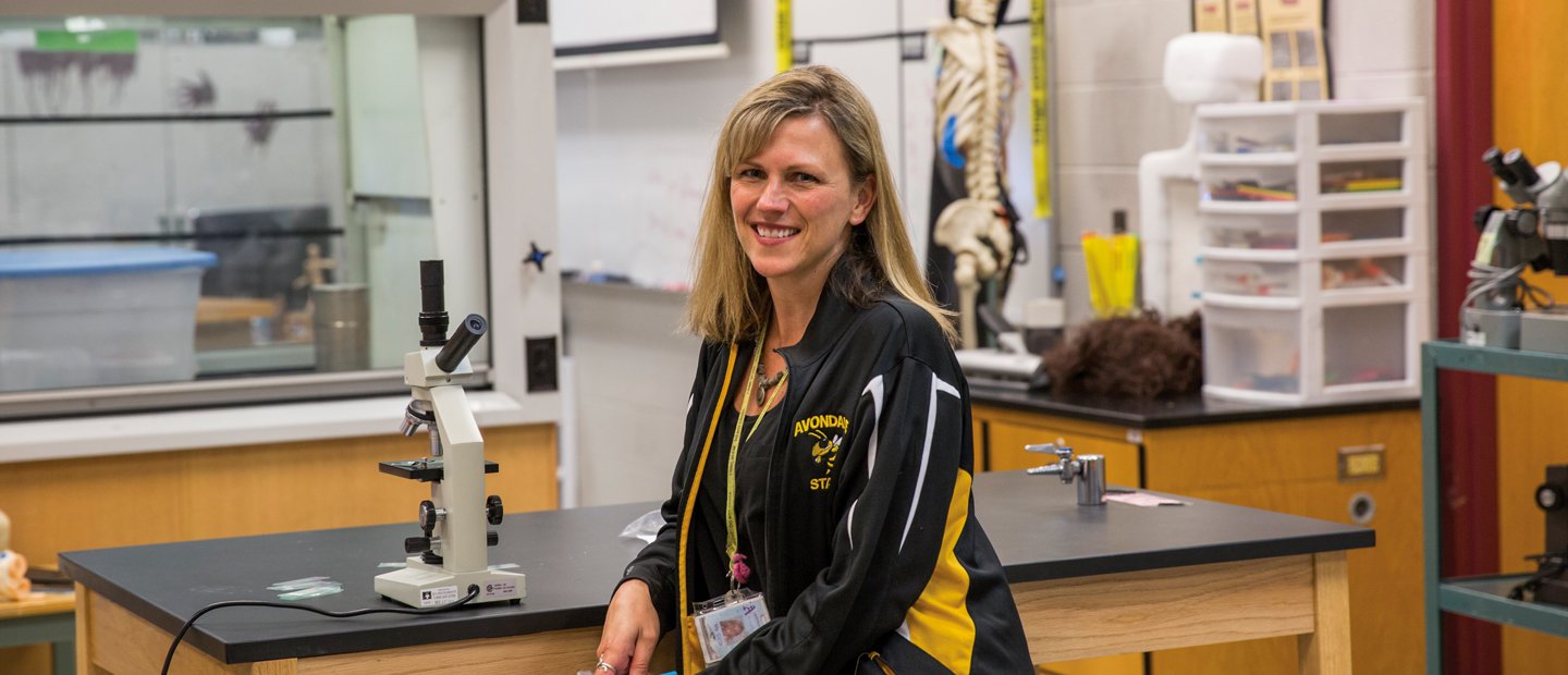 A teacher in an athletic jacket seated at a lab table with a microscope on it.