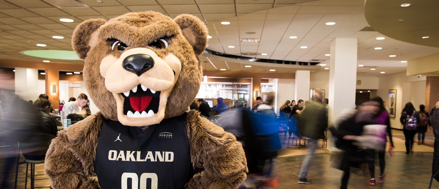 The Grizz bear wearing a jersey that says Oakland on it, with people walking through the room behind him