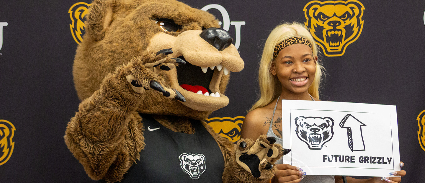 A new Oakland University student holding a Future Grizzly sign and posing with the Grizz mascot