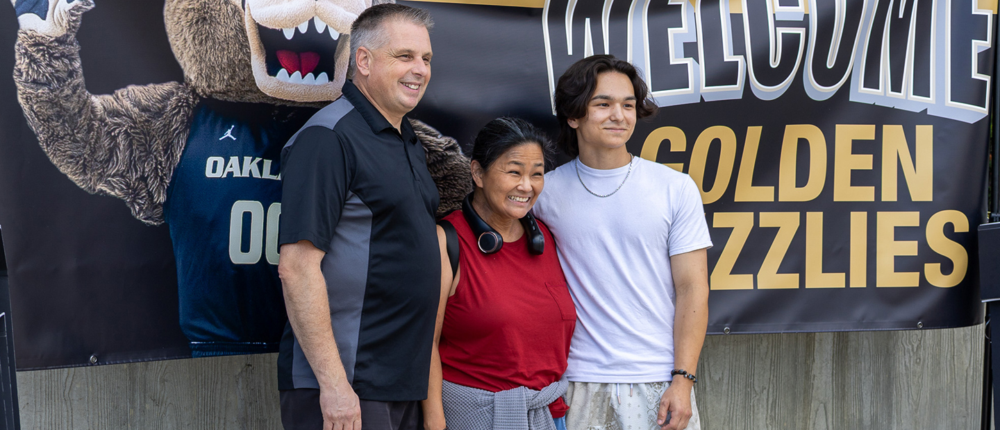 A new student and his parents in front of a Welcome Golden Grizzlies banner